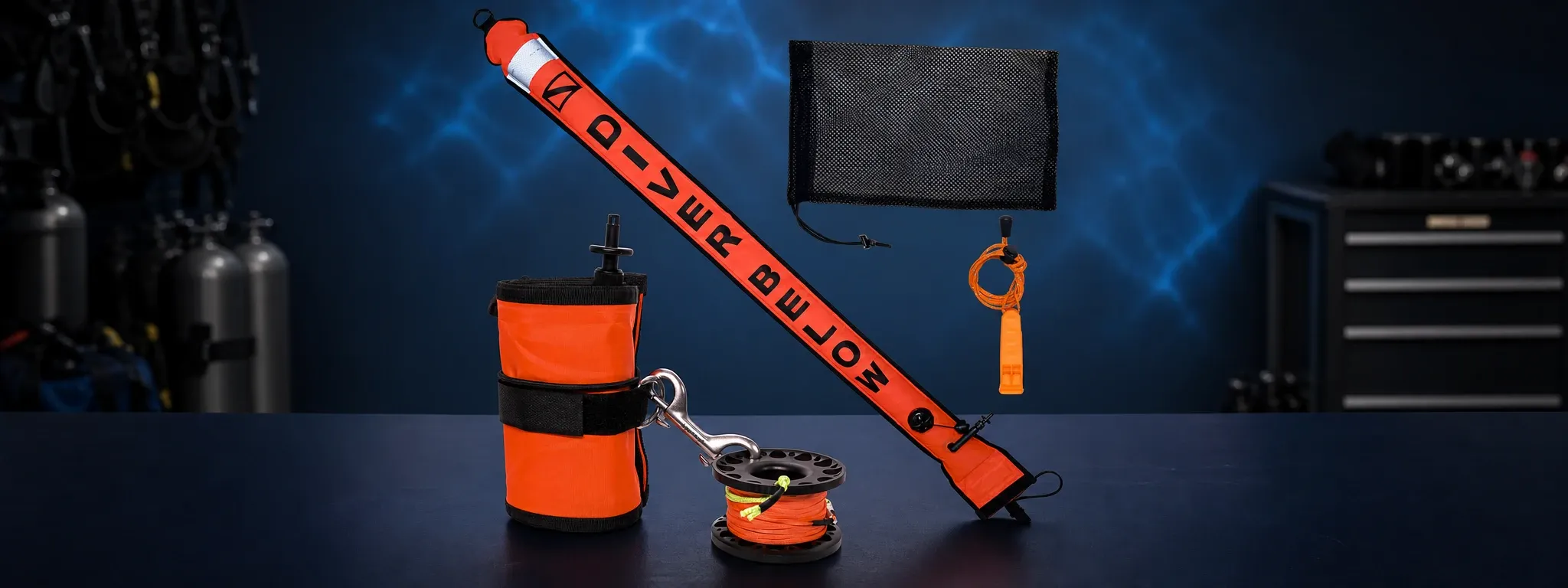 Flat lay of the all-inclusive surface safety kit for UK scuba divers, featuring an unrolled bright orange DSMB with black text, a spool with orange line, a marine-grade stainless steel double-ended clip attached to an orange pouch, an orange whistle with cord, and a black mesh bag, all set against a dark blue background with subtle water light reflections and scuba tanks in the background.